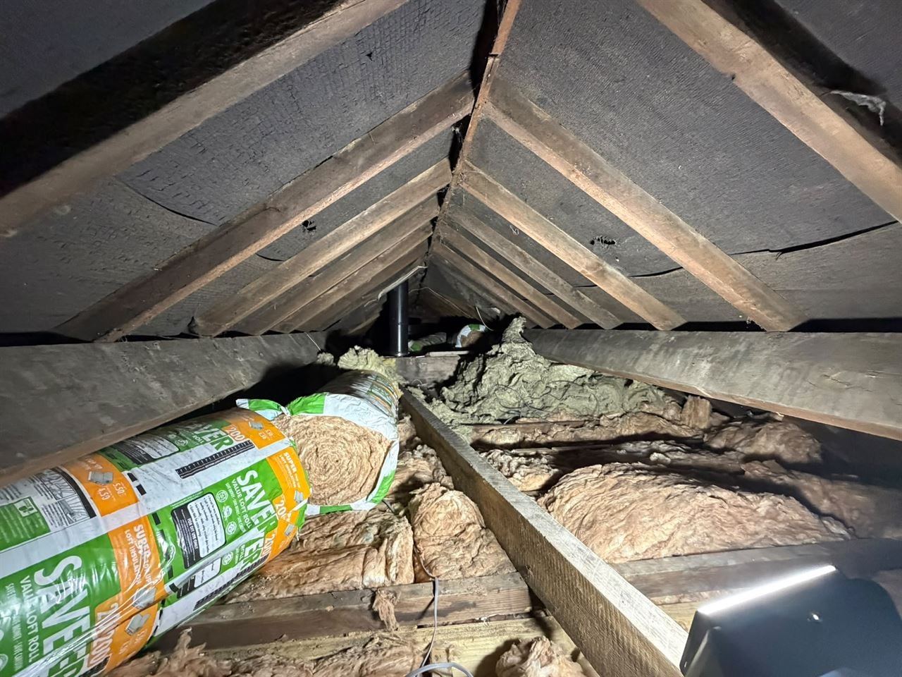 Dark loft roof with old fibreglass insulation between the joists, before it was removed, and a roll of loft insulation resting on the timbers