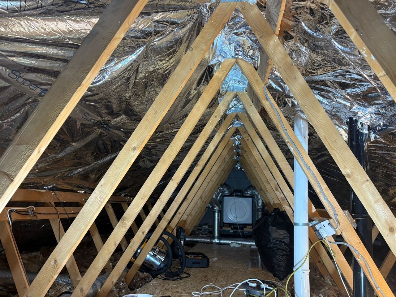 Inside a loft: timber roof trusses with SuperFOIL insulation lining the roof, and a white unit with silver ducting at the far end